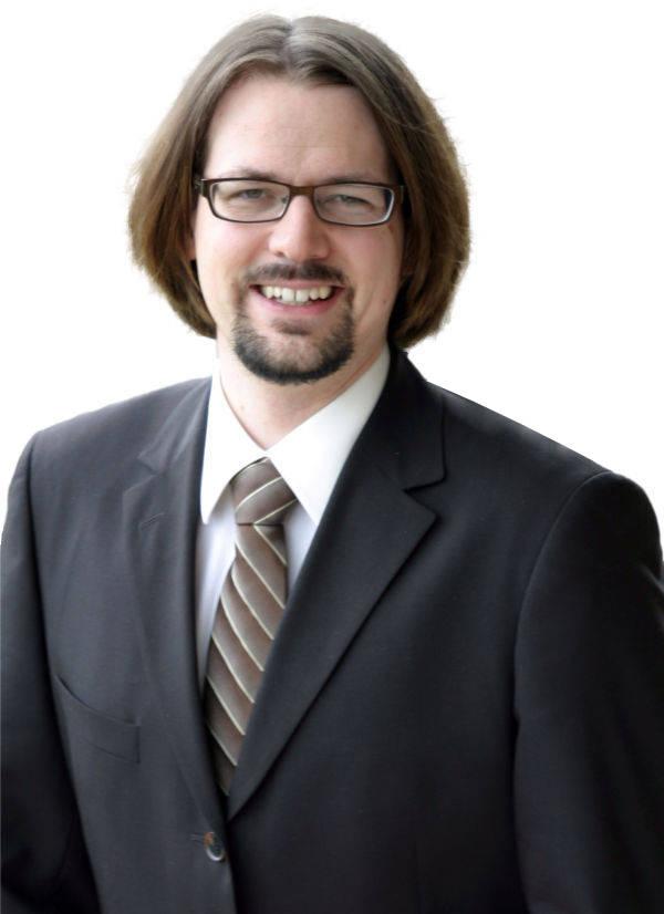 Holger Wöltje, expert in productive work with Office 365: a man with shoulder-length hair, glasses, a pointed goatee and a suit, smiling at the camera.